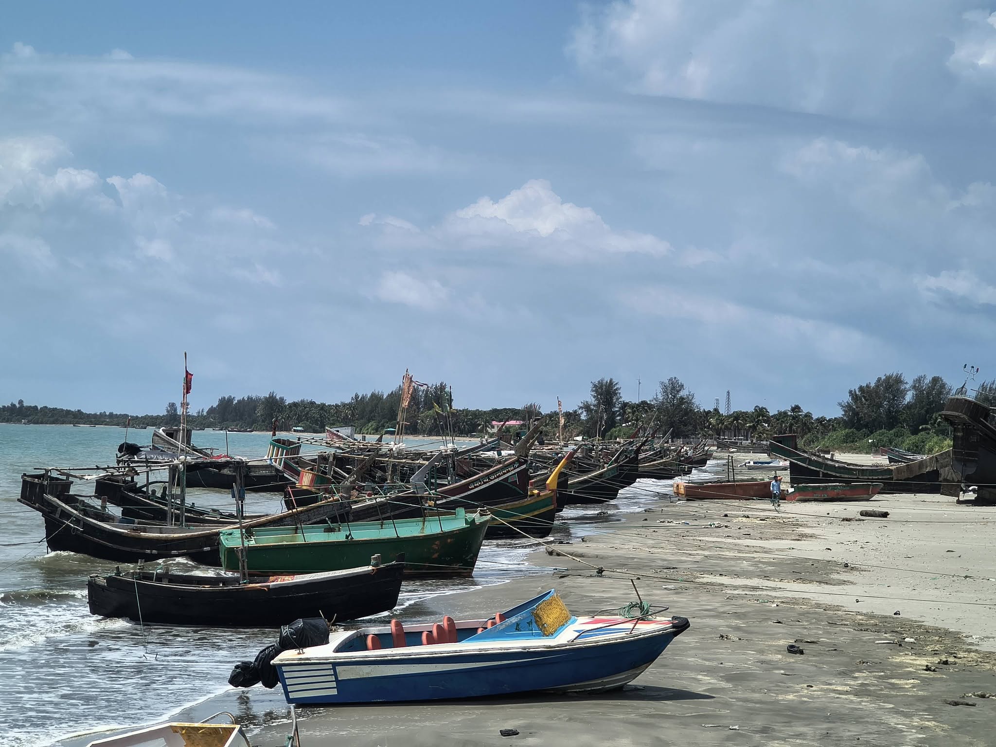 সেন্টমার্টিনের সৈকতে এই দৃশ্যটি কি অসাধারণ! সারি সারি কাঠের ট্রলার, তাতে উড়ছে কালো পতাকা—যেন এক নতুন অ্যাডভেঞ্চারের প্রস্তুতি চলছে। তটরেখায় মানুষের কর্মব্যস্ততা আর সমুদ্রের শান্ত পরিবেশ, প্রকৃতির এক চমৎকার মেলবন্ধন। ছবিতে ‘রিফা’ ট্রলারটির নামও পরিষ্কার দেখা যাচ্ছে, যা দৃশ্যটিতে একটি ব্যক্তিগত স্পর্শ যোগ করেছে।